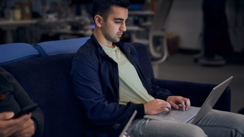 Lightly bearded man working on a laptop while sitting next to another person on a dark blue couch